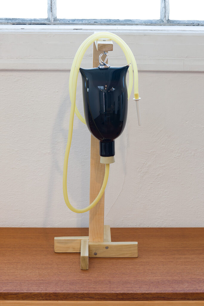 Image of a black blown glass enema bag with a tube hanging from the bottom. The black bag is hanging on a wooden stand.