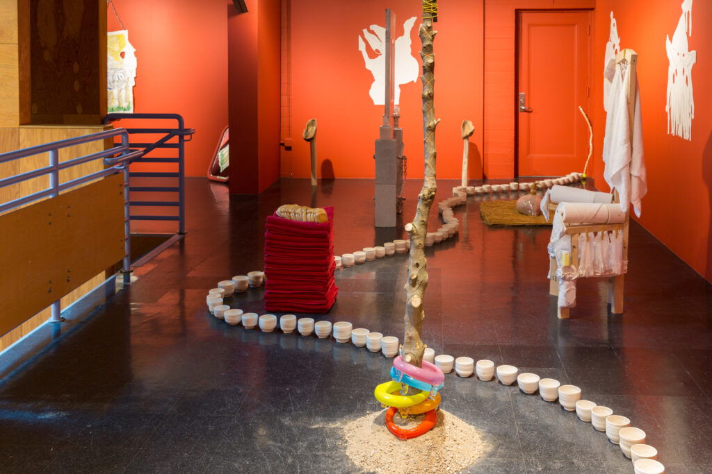 Image of a large room filled with various art objects. In the forefront is a branch of a tree standing up from a pile of sand with colorful glass rings around the base. There is a chain of white ceramic cups snaking through the room on the floor. A stack of red towels with a loaf of Wonder Bread on top. There is a thrown like chair with an enema bag of milk hanging from the top.