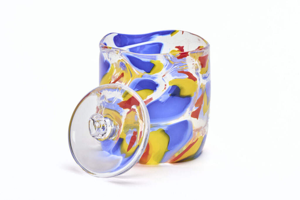 Large blown glass cylinder with blue, yellow and red wavy blotches of color throughout. Leaning against the side is a solid hot sculpted glass weight with a ring on the top as a handle