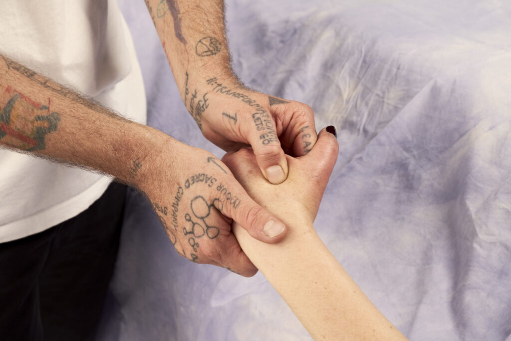 A person with tan skin and tattoos giving a person with pale soft skin and painted black fingernails a hand massage.