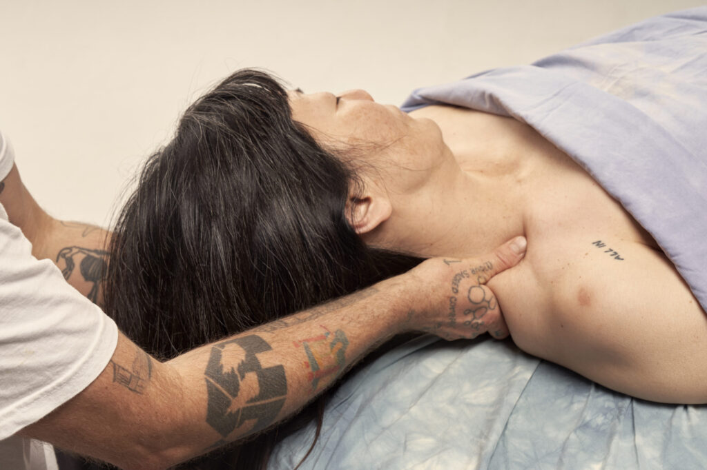Image a person of Korean decent with pale soft skin and long black hair getting a massage on her neck from a person with tan skin and tattoos. The thumb is pressing into the space just above the collar bone.