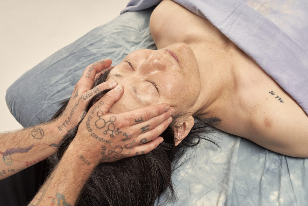 A Person of Korean decent with pale soft skin and black hair, eyes closed, getting a head massage from a person with tan skin and tattoos. The thumbs are gently resting on the forehead and the fingers are applying gentle pressure to the temples.