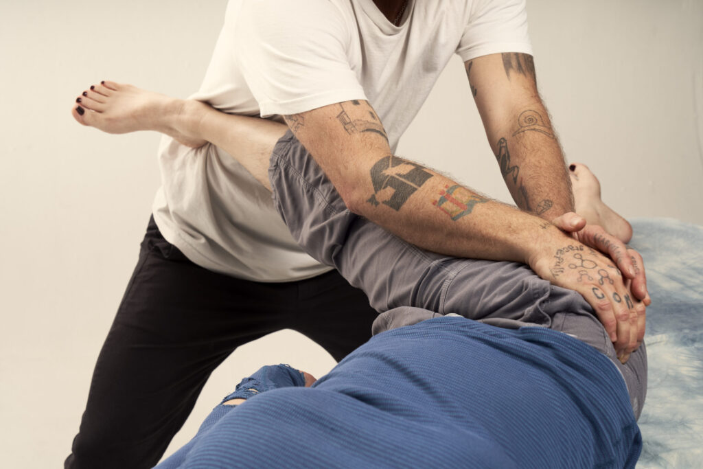 Image of a person with pale soft skin and painted black toe nails wearing grey pants and a blue shirt getting an assisted hip stretch from a tan person with tattoos wearing a white t-shirt and black pants.