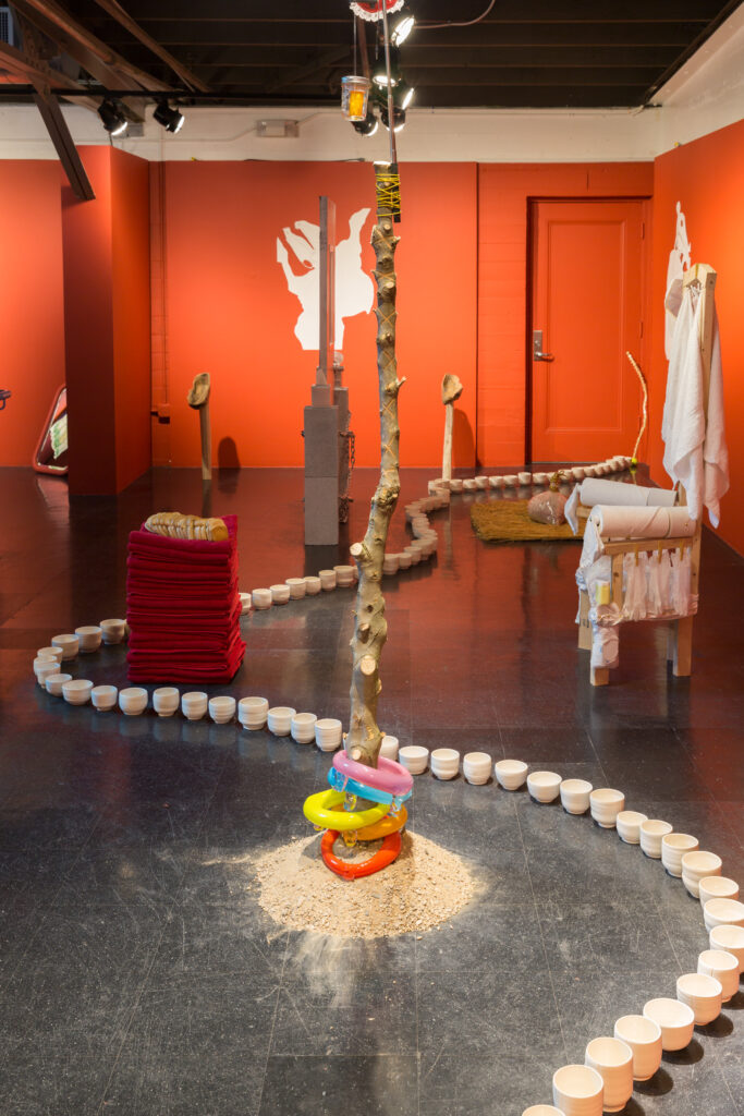 Image of a large room filled with various art objects. In the forefront is a branch of a tree standing up from a pile of sand with colorful glass rings around the base. There is a chain of white ceramic cups snaking through the room on the floor. A stack of red towels with a loaf of Wonder Bread on top. There is a thrown like chair with an enema bag of milk hanging from the top.
