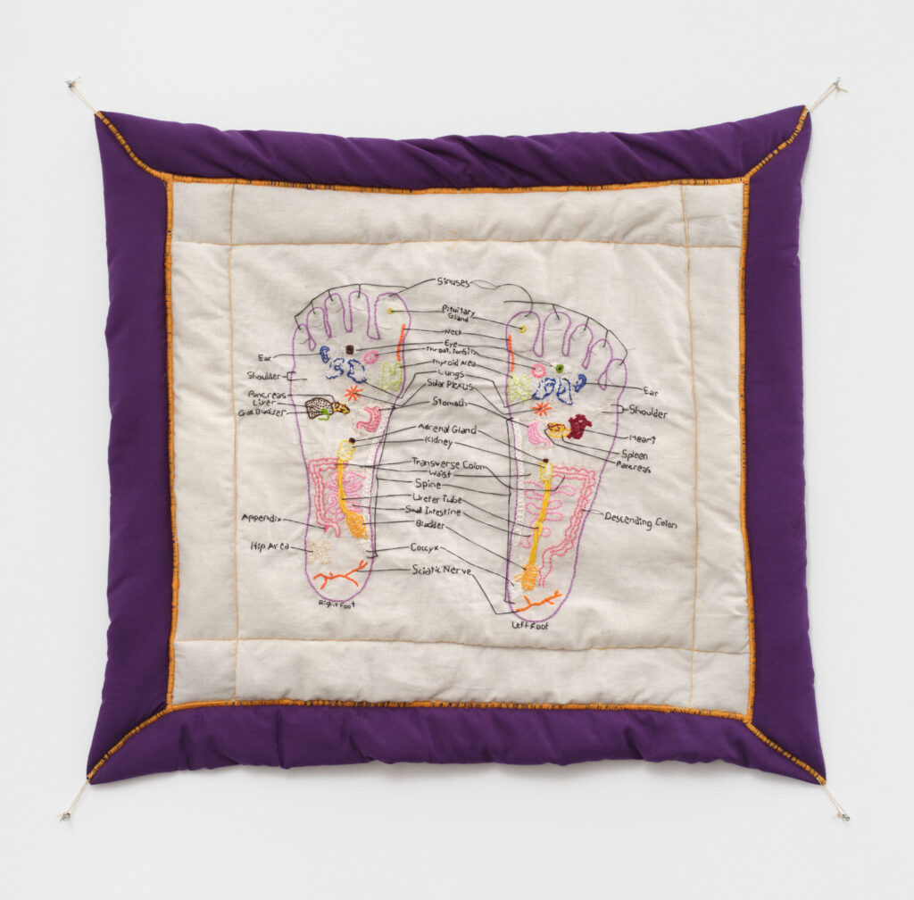 A colorful embroidered diagram of a reflexology chart of the foot. It is on a quilted white background with a purple frame and gold thread accenting the outline.