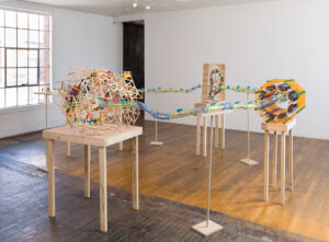 Image of a several sculptures on table tops that are a fantastical representation of the Large Hadron Collider. It is made of hamster tubes, KNex, popsicle sticks and foam. It as a playful feel with a lot of bright colors.