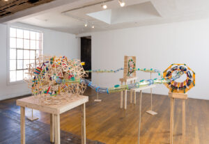 Image of a several sculptures on table tops that are a fantastical representation of the Large Hadron Collider. It is made of hamster tubes, KNex, popsicle sticks and foam. It as a playful feel with a lot of bright colors.