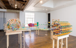 Image of a several sculptures on table tops that are a fantastical representation of the Large Hadron Collider. It is made of hamster tubes, KNex, popsicle sticks and foam. It as a playful feel with a lot of bright colors.