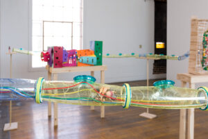 Detail image of the JLS sculpture. It shows two action figures inside a hamster tube with colorful wires running the length of the tubes. The figures are smashed up and in a bundle together.