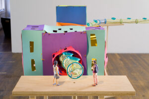Image of two small figurines, much like action figures standing on a platform in front of a large sculpture the is a fantastical representation of the large hadron collider. One is of Korean descent and is wearing a skirt with long black hair. The other is whiter, non-binary, wearing pink pants and pink shirt with a backpack on. They look as though they are waiting to go in.