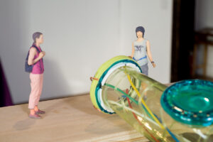 Image of two small figurines, much like action figures standing on a platform in front of a large sculpture the is a fantastical representation of the large hadron collider. One is of Korean descent and is wearing a skirt with long black hair. The other is whiter, non-binary, wearing pink pants and pink shirt with a backpack on. They look as though they are waiting to go in.