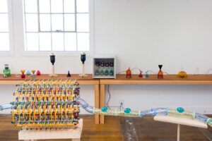 Image of a long wooden table against a wall with various colorful blown glass vessels and a small refrigerator. It is reminiscent of a science lab. In the foreground you can see parts of the JLS sculpture.