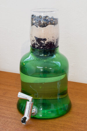 Image of a blown glass green vessel with a spout on it. Inside the vessel is water. On top of the vessel is blown glass funnel with layers of different kinds of rocks and sand. It is a filter.