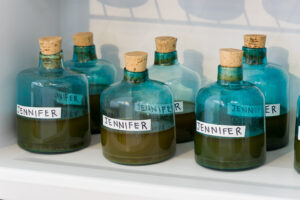 Image of a shelf inside a small fridge stacked with blue blown glass bottles with a brown liquid inside of each of the vessels. There is a label on each bottle that reads Jennifer.