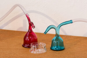 Image of two blown glass vessels, one is pink and one is blue with two different bottle necks on each vessel. One of the bottle necks is attached to a tube and the other on both vessels is pouring into a small blown clear glass bowl.