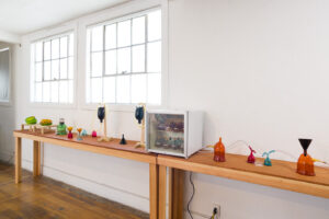 Image of a long wooden table against a wall with various colorful blown glass vessels and a small refrigerator. It is reminiscent of a science lab.