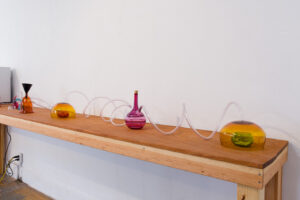 Image of a long wooden table against a wall with various colorful blown glass vessels and tubes connecting the vessels. It is reminiscent of a science lab.