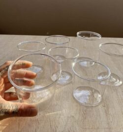 seven clear blown glass cups with a white hand reaching into the frame and picking one of them up.