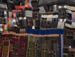 Earth 2022 This is a detail of the Earth quilt. A large landscape pillowy quilt. The quilt is made out of Harris tweed scraps of various color and shape. It is sewn together to look like an abstracted areal view of a landscape. The quilt is overstuffed and pinched in different places giving it a pillowy thickness. The back of the quilt is an orange satin material that can be seen from the front around the edges. It is hanging on a white wall stretched from its corners. There is a heaviness and weight to it that makes it sag.
