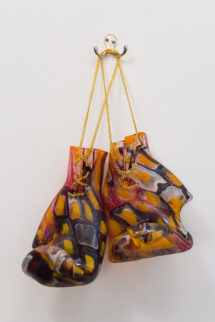 A pair of blown glass boxing gloves with abstract patterns of red, yellow and black squares throughout. They have gold chain laces and are hanging from a hook on the wall.