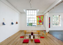 Installation view of the exhibition The Act of Growing Up: Emanations of a Soul Nerve Revival. It shows a bench with two stations to wash feet, glass boxing gloves hanging on the left wall, a large window on the back wall, and a hanging flag like tapestry with a girl eating ice cream.