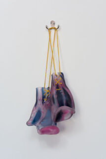 A pair of blown glass boxing gloves with abstract patterns of pink, dark blue and clear circles throughout. They have gold chain laces and are hanging from a hook on the wall.