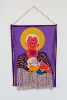 A textile banner with an image of an older woman holding a baby. The baby is sucking on her finger and she has an ecstatic look on her face. There is a gold halo encircling her head. The background is dark purple. There are strings of pastel beads hanging from the bottom of the tapestry. The tapestry hangs from a gold rope on the wall.