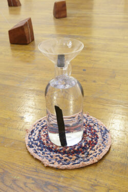 Water Station
2015 Image of a clear blown glass bottle that is sitting on a pastel colored round rug on the floor. The bottle has a blown glass funnel that is resting inside the neck of the bottle. Inside the bottle is water and sticks of charcoal are floating in the water.