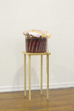 Sauerkraut House
2014-15 Image of a blown glass cylinder on a small wooden stool. The cylinder has brown stripes on it. There is fermenting cabbage in the vessel and there is a glass weight on top of the fermenting vegetables. There is a sewn fabric cloth covering the top of the vessel.