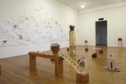Installation View
2015 Installation view of the exhibition Oh Be Gentle. It shows a pyramid of drawings on the wall in the background, with a bench holding a vessel on it in the foreground. There is a cabinet like structure in the center of the room holding a glass vessel on top of it. Wood chunks encircling it. You can see the entrance to the room.