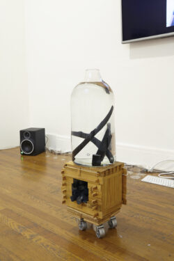 Water Cart
2014-15 Image of a large clear blown glass bottle sitting on top of a small wooden square cart with wheels. The cart holds a stack of sticks of charcoal. In the vessel is water with several sticks of charcoal floating in the water.