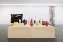 Let Me Touch You 2023 Installation view of the exhibition Let Me Touch You. A large wooden platform in the foreground with fifteen uniquely shaped ceramic vessels in primary colors on top. In the background on the wall are two large quilts and several glass sculptures.