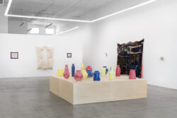 Let Me Touch You 2023 Installation view of Let Me Touch You. A large wooden platform in the foreground with fifteen uniquely shaped ceramic vessels in primary colors on top. In the background on the walls are large quilts and several glass sculptures.