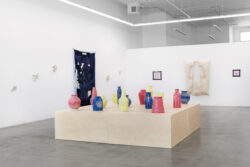 Let Me Touch You 2023 Installation view of Let Me Touch You. A large wooden platform in the foreground with fifteen uniquely shaped ceramic vessels in primary colors on top. In the background on the walls are large quilts and several glass sculptures.