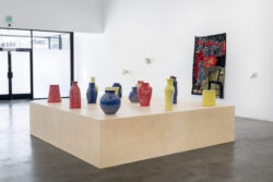 Let Me Touch You 2023 Installation view of the exhibition Let Me Touch You. A large wooden platform in the foreground with fifteen uniquely shaped ceramic vessels in primary colors on top. In the background on the wall is a large quilt and glass sculptures. You can see the doors of the gallery.