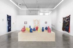 Let Me Touch You 2023 Installation view of the exhibition Let Me Touch You. A large wooden platform in the foreground with fifteen uniquely shaped ceramic vessels in primary colors on top. In the background on the walls are several large quilts and glass sculptures.