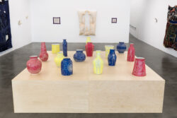 Let Me Touch You 2023 Installation view of Let Me Touch You. A large wooden platform in the foreground with fifteen uniquely shaped ceramic vessels in primary colors on top. In the background on the wall are three quilts.