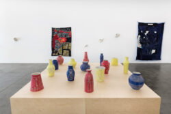 Let Me Touch You 2023 Installation view of Let Me Touch You. A large wooden platform in the foreground with fifteen uniquely shaped ceramic vessels in primary colors on top. In the background on the wall are two large quilts and several glass sculptures.
