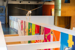Image of a flying dog skeleton with attached hot sculpted glass orange, white and black, angel wings. Below you can see a stairwell with colorful banners hanging on the walls.