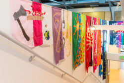 Image of a stairwell with colorful banners hanging from the wall telling the life story through sewn pictures, much like a comic book, of the dog Mr. Snuggles.