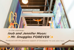 Image looking up the stairwell of the exhibition Mr. Snuggles Forever. On all the walls are banners portraying different stories from the dog's life. In the foreground, "laub and Jennifer Moon: Mr. Snuggles Forever" is printed in black onto a white post that extends the width of the stairwell.