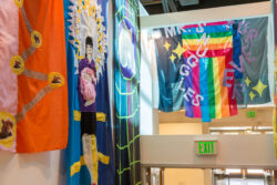 Image of a stairwell with colorful banners hanging from the wall telling the life story through sewn pictures, much like a comic book, of the dog Mr. Snuggles.