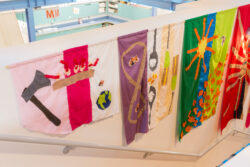 Image of a stairwell with colorful banners hanging from the wall telling the life story through sewn pictures, much like a comic book, of the dog Mr. Snuggles.