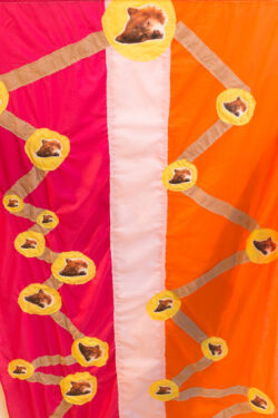 A detailed image of one of the textiles from the exhibition Mr. Snuggles forever. This tapestry portrays small molecular yellow circles with a print of Mr. Snuggles' face on top them. The circles are all connected with brown lines. The background is pink and orange.