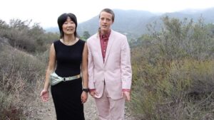 Video still of two people walking down a path talking to the camera. One person is of Korean descent with long black hair and wearing a black dress with a green fanny pack. The other person is white and non binary and is wearing a pink suit.