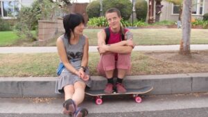 Video still of two people, one with long black hair of Korean descent with a skirt on and her legs stretched out in front of her and the other white and non-binary wearing all pink, with their feet on a skateboard. They are sitting next to each other on the edge of a sidewalk, talking.